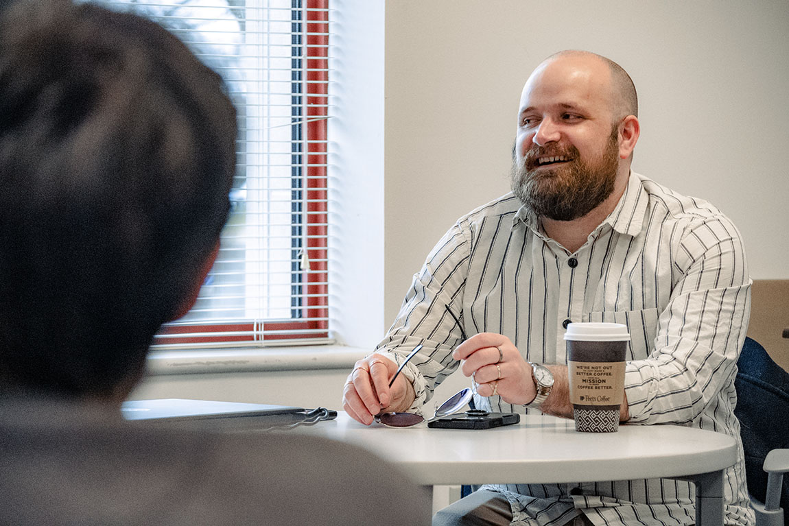 A man with a brown beard smiles and looks to his right while sitting at a table